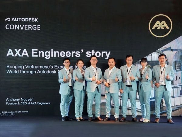 A group of seven people in matching light blue suits stands in front of a large screen displaying AXA Engineers Story and Bringing Vietnamese expertise to the world through Autodesk Converge. Showcasing their civil engineering services, they all give a thumbs-up, smiling for the camera.