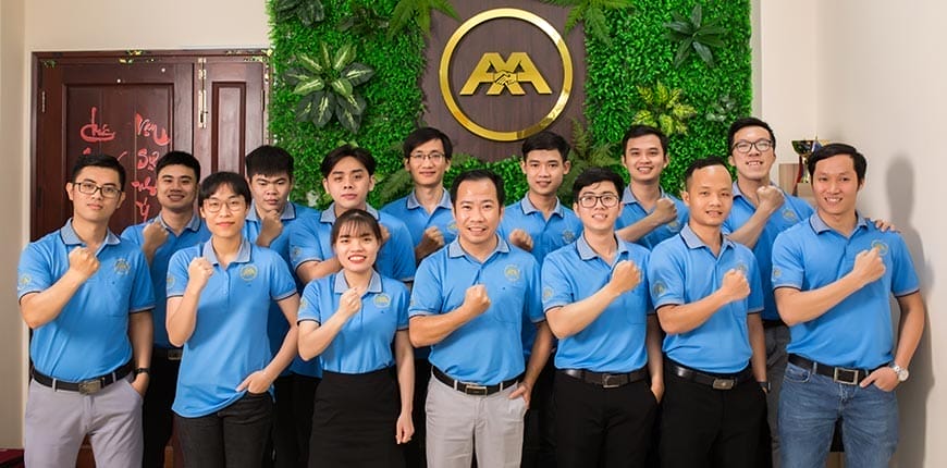 A group of people wearing matching blue polo shirts stand indoors, smiling and raising one fist in a gesture of solidarity. They are positioned in front of a green wall with a logo featuring the letters MA, making a bold statement for branding.
