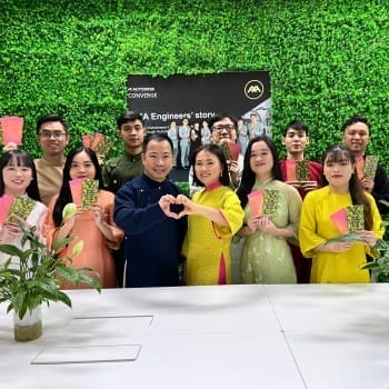 A group of people stands smiling in front of a lush green wall, holding colorful envelopes. Two people in the front center make a heart shape with their hands. Behind them, a poster showcases civil engineering outsourcing, adding a professional touch to the joyous scene.