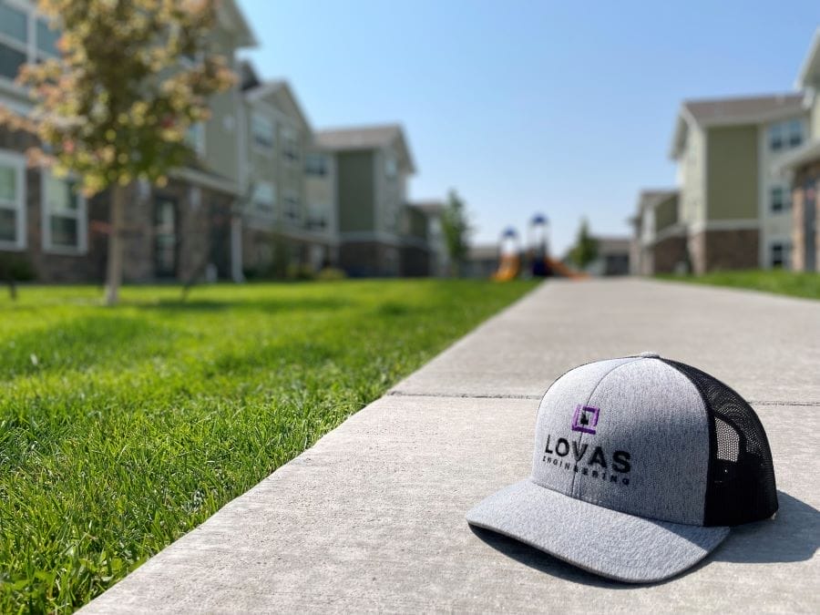 A gray and black baseball cap with the AXA Engineers logo sits on a concrete pathway in a residential area. The pathway is flanked by grass and trees, leading toward modern apartment buildings, with a blurred view of a playground in the distance.