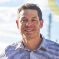 A smiling man in a checkered shirt stands outdoors, embodying the spirit of civil engineering amidst a backdrop of blue skies and scattered clouds.