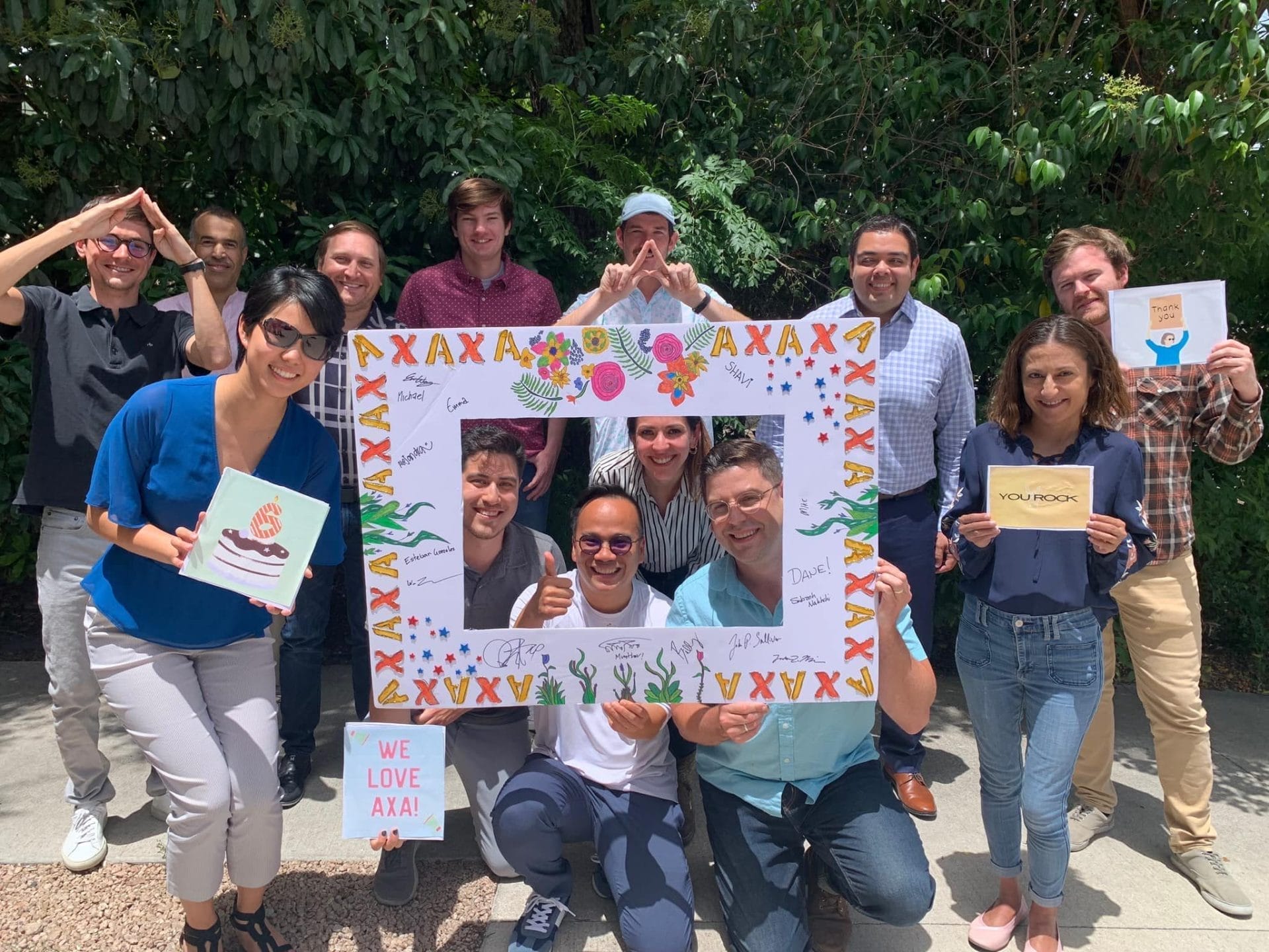 A group of people, possibly AXA Engineers, smiling outdoors, hold artwork and signs. One sign reads WE LOVE AXA Engineers and another YOU ROCK. They gather around a colorful frame with signatures. Lush greenery sets a vibrant backdrop for this scene of pioneering progress.