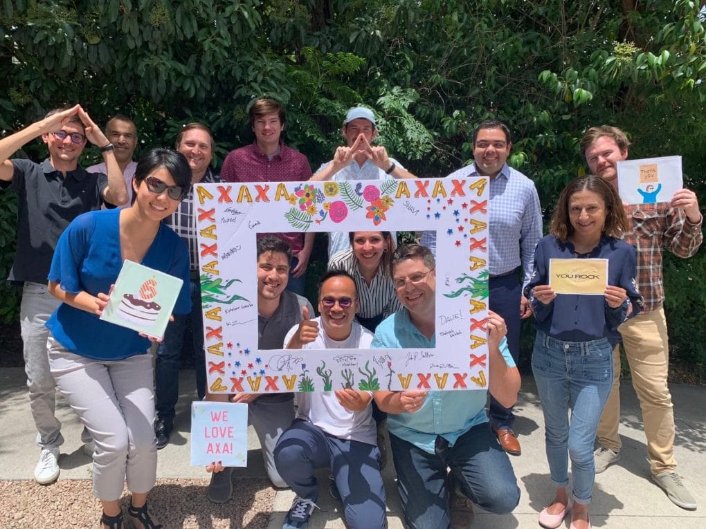 Pioneering Progress Together: Civilitude’s Journey with AXA Engineers 2 A group of people, possibly AXA Engineers, smiling outdoors, hold artwork and signs. One sign reads WE LOVE AXA and another YOU ROCK. They gather around a colorful frame with signatures. Lush greenery sets a vibrant backdrop for this scene of pioneering progress.