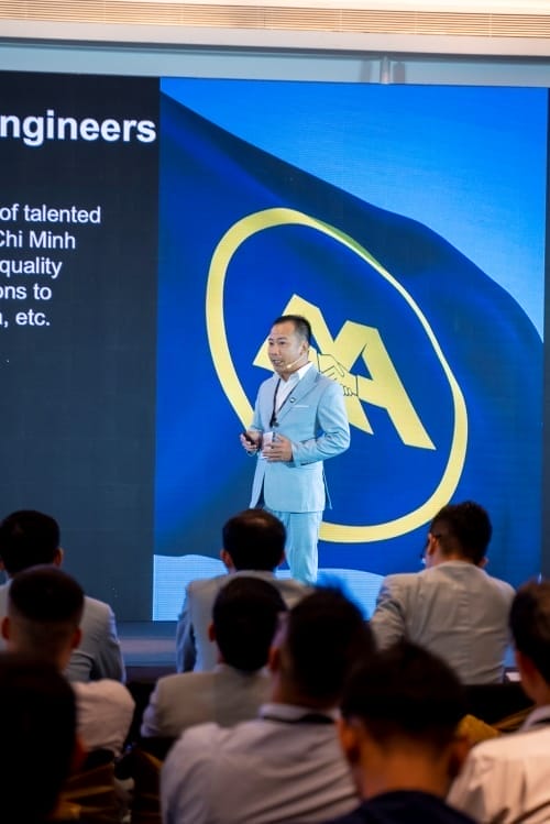 A man in a light blue suit representing AXA Engineers Services is speaking on stage at a conference. Behind him, a large screen displays text alongside a blue flag with a yellow emblem, capturing the audiences attention as they sit before the stage.