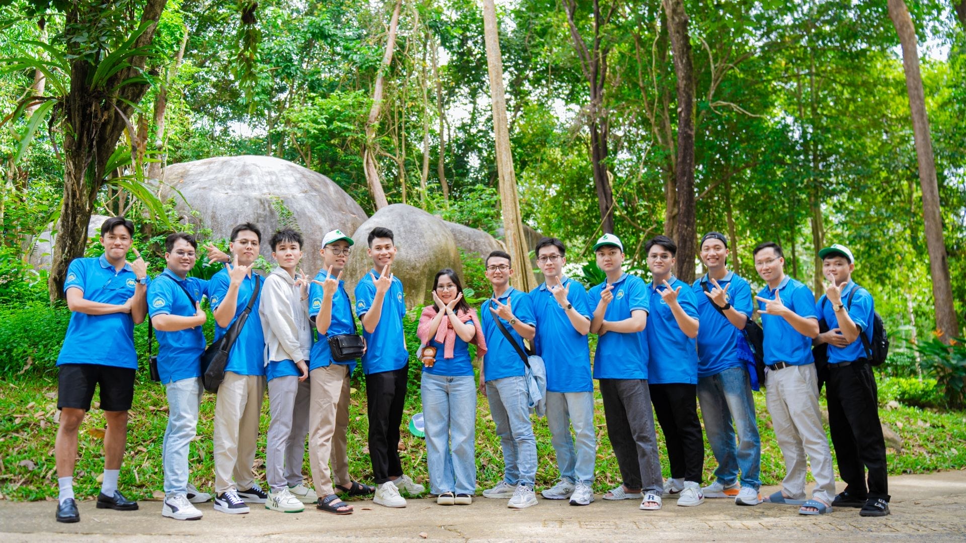 A group of people in blue shirts stands in a line outdoors in a lush green forest setting, showcasing the spirit of AXA Engineerss team building. They are posing with various hand gestures, smiling at the camera.