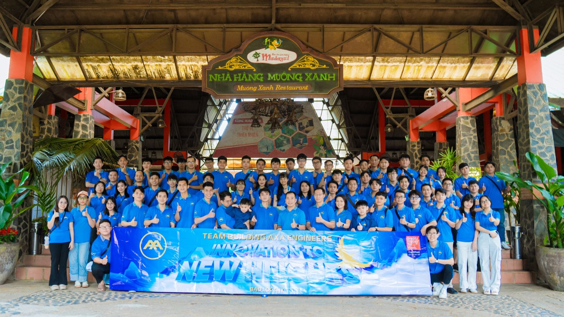A large group of people in matching blue shirts pose for a group photo in front of a building with a sign that reads Nhà Hàng Mường Xanh. They proudly hold a blue banner that says Innovation - New Milestones, marking their successful team building event for AXA Engineers 2024.