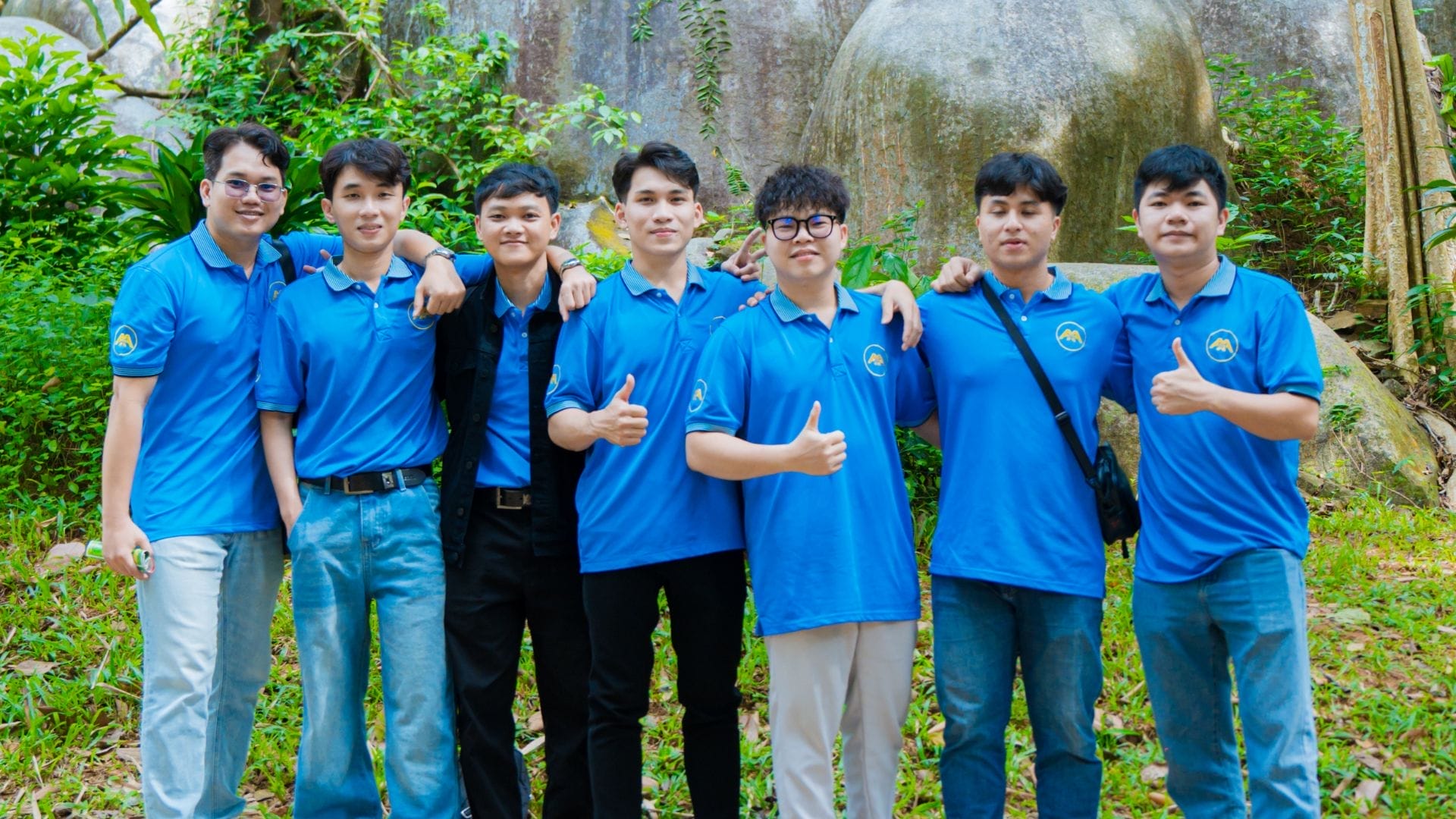 Seven young men stand in line outdoors, proudly wearing matching AXA Engineers blue polo shirts with a logo. With thumbs up and arms around each other, they smile against a backdrop of greenery and rocks, showcasing the spirit of team building and innovation.