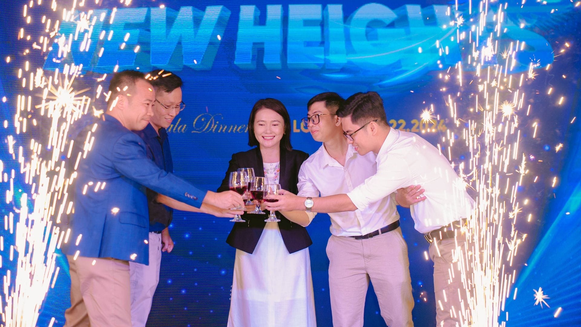 Five people are gathered, raising glasses in a toast as fireworks sparkle around them. Standing before a blue backdrop with New Heights in large text, they celebrate innovation and team building at AXA Engineers.