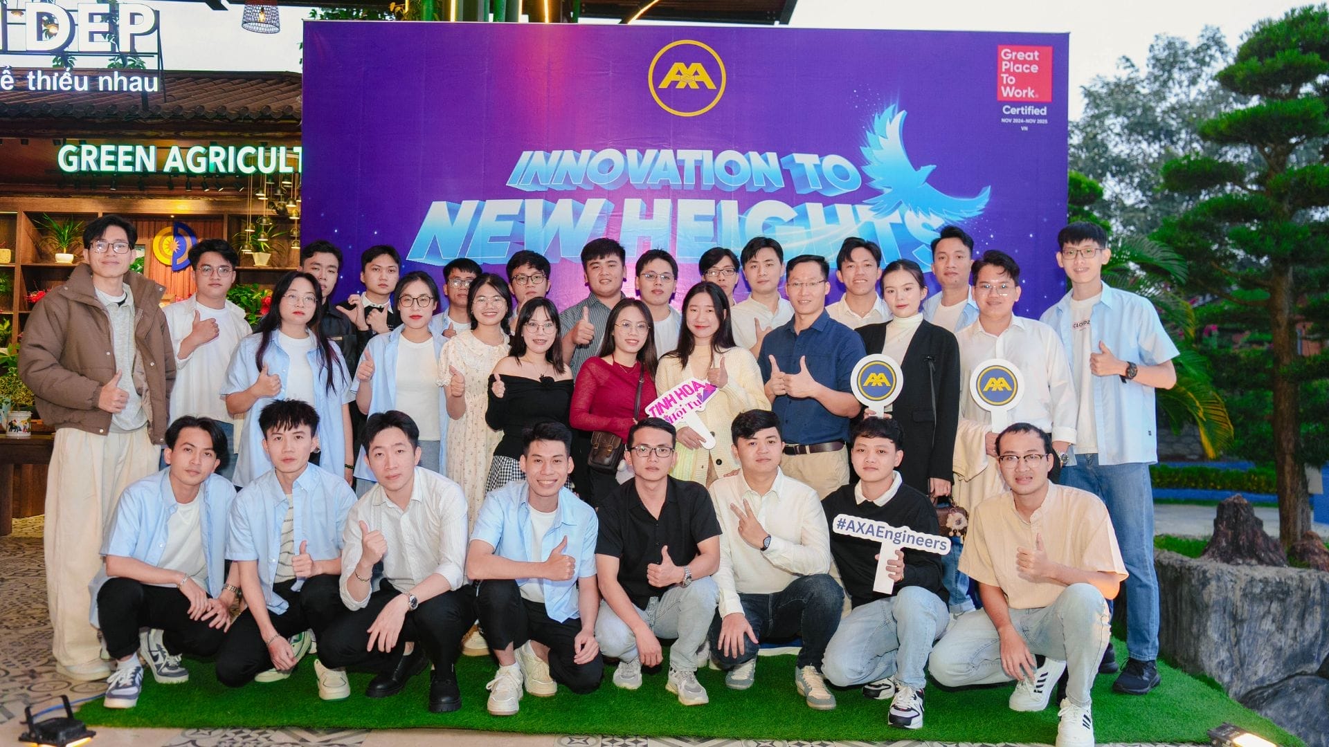 A large group of people from AXA Engineers gather outside, smiling broadly in front of a banner that reads Innovation to New Heights. Some hold signs with hashtags. Clad in casual attire, they celebrate team building amid greenery as the evening sky envelopes them.