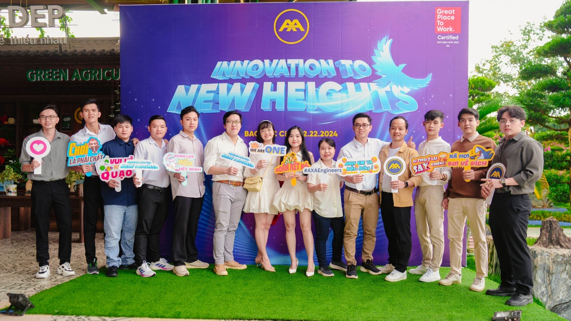 A group of people stands on the grass, proudly holding colorful signs with messages like Innovation to New Heights and a backdrop that reads Great Place to Work Certified, dated 8.22.2024. This vibrant scene captures the spirit of team building and celebrates AXA Engineers 2024s commitment to innovation.