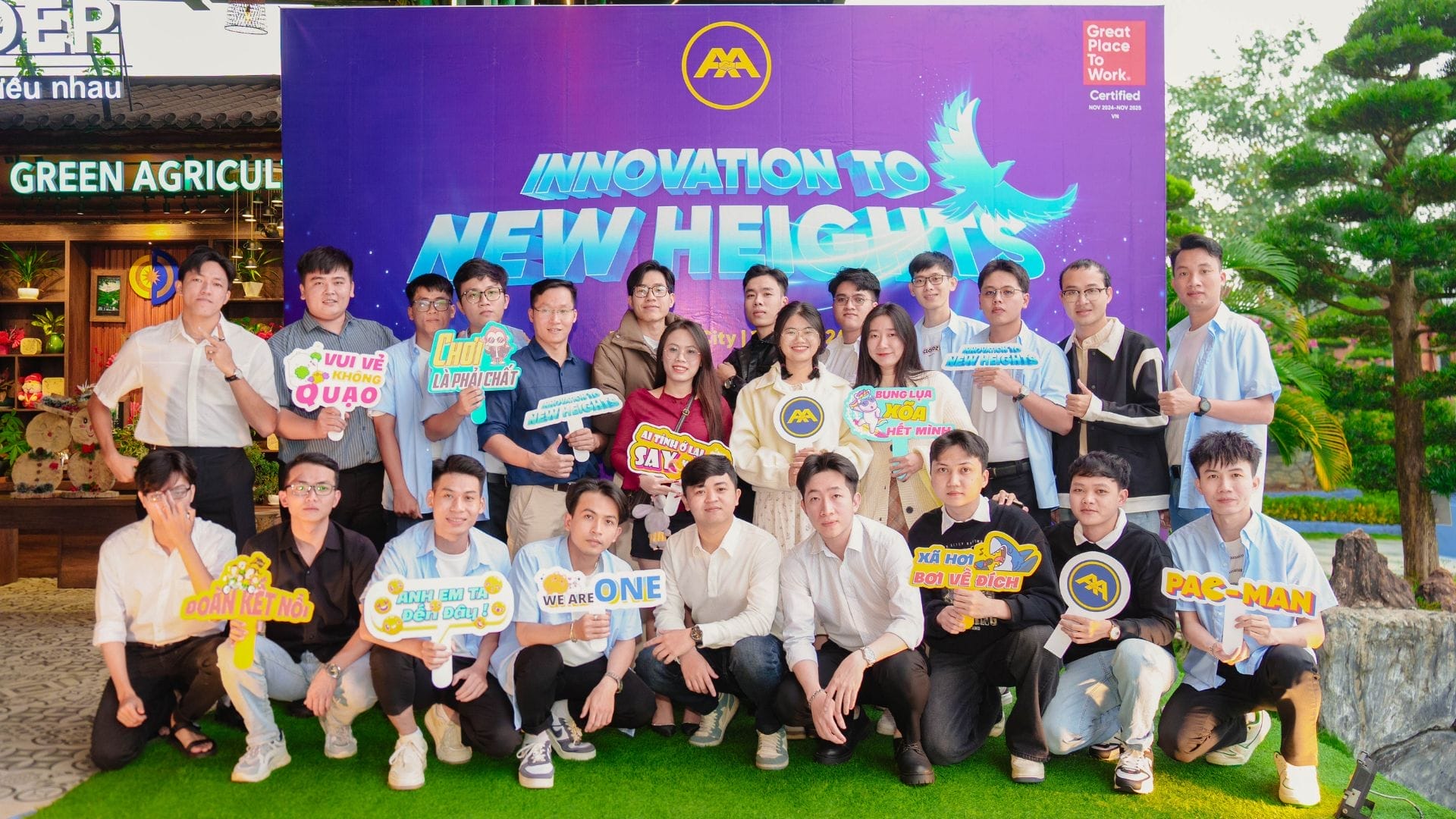 A group of people pose in front of a colorful backdrop with the text Innovation to New Heights, embodying AXA Engineerss spirit. Theyre holding various signs with cheerful messages. The setting appears festive and outdoors, adorned with green plants and decorative elements.