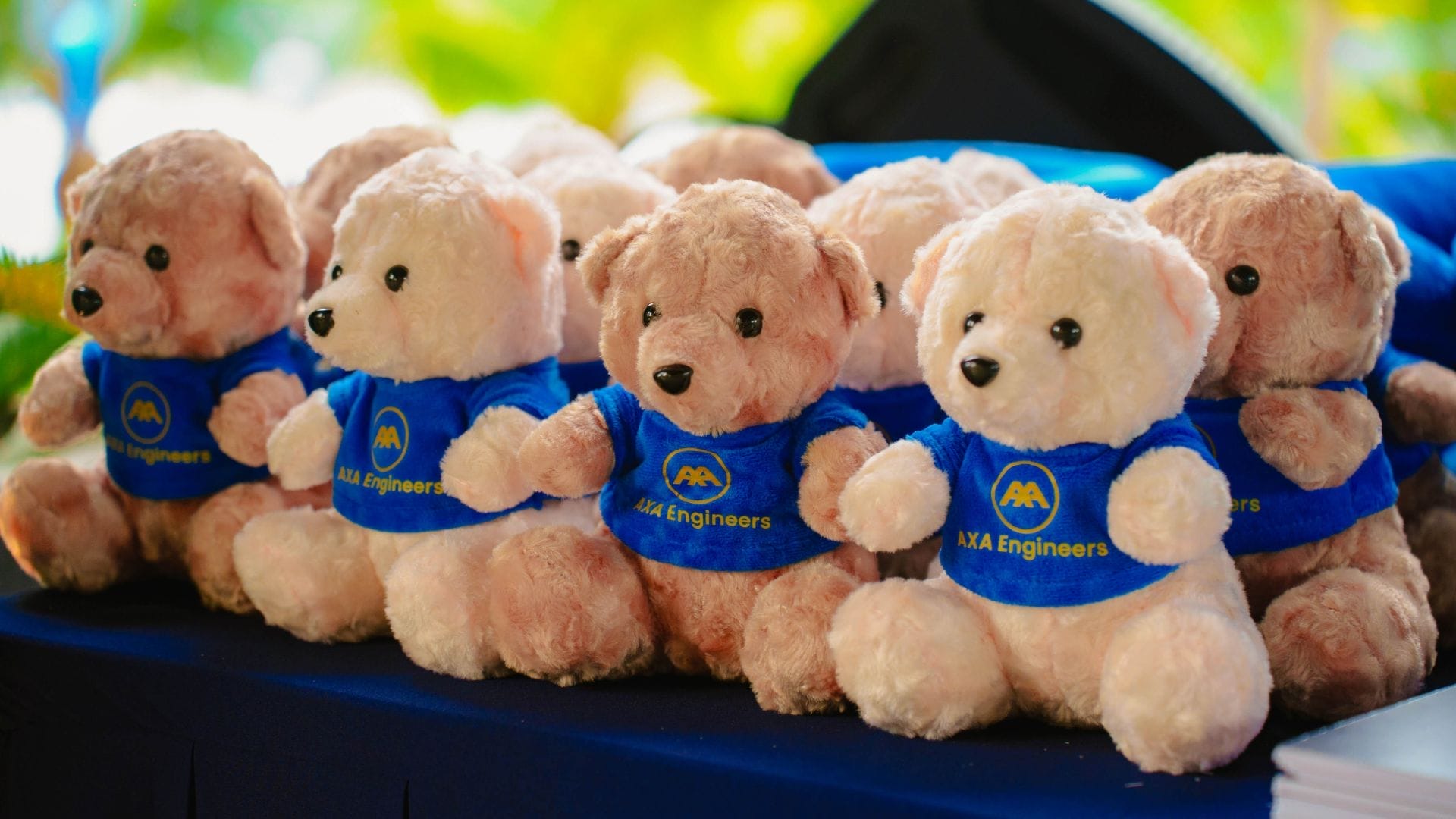 A row of identical teddy bears wearing blue shirts with the AAA Engineers logo sits on a dark surface, symbolizing team building. Bright green foliage blurs in the background, echoing innovation.