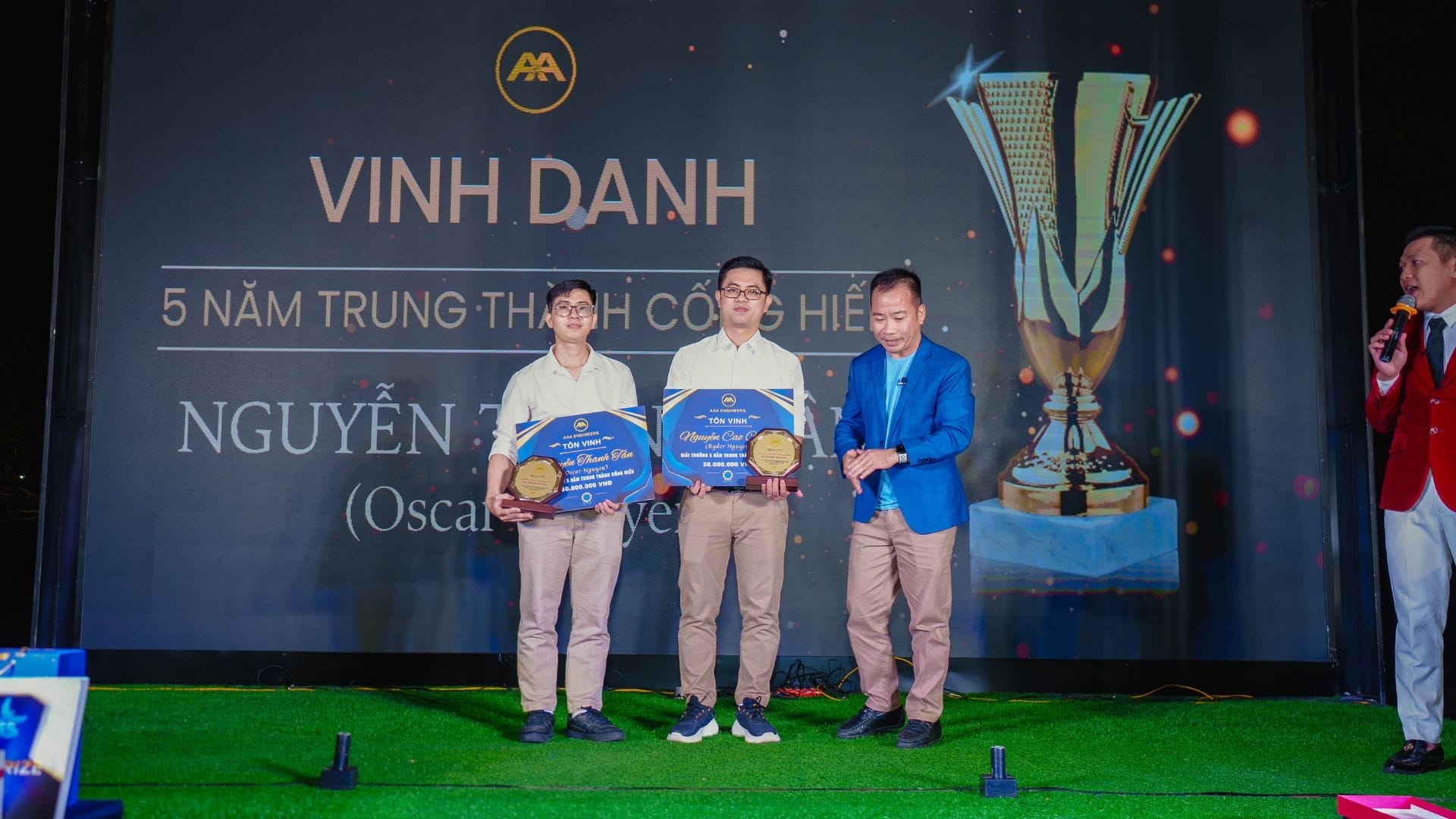 Three men stand on a stage holding awards, embodying innovation. The background displays a large trophy image and Vietnamese text. One man speaks into a microphone, while two others, holding plaques, pose for a photo. They are dressed in casual and semi-formal attire.