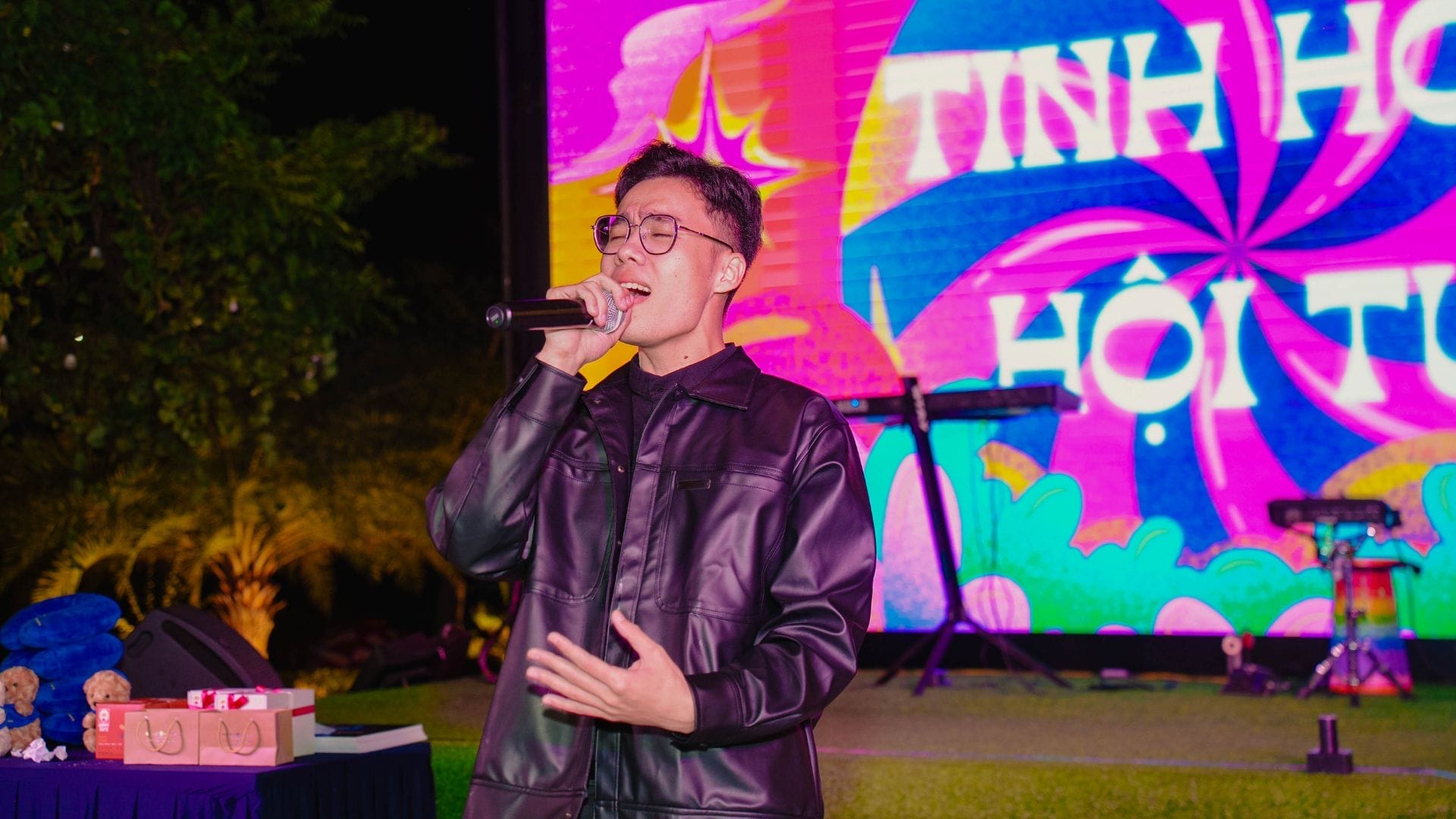 A person wearing glasses and a dark jacket is passionately singing into a microphone on an outdoor stage. A colorful psychedelic background displays Vietnamese text, reflecting the spirit of innovation. Nearby, a keyboard and blue teddy bear add charm to this vibrant scene.
