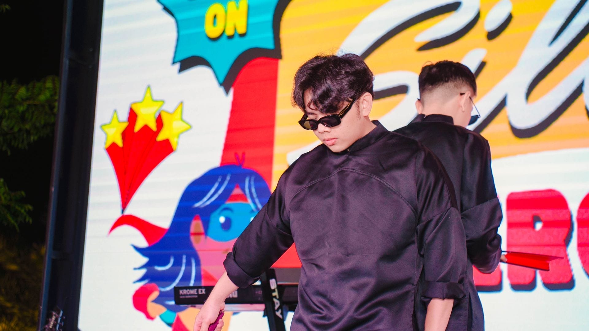 A person in a black outfit and sunglasses performing on stage, set against a vibrant animated backdrop of stars and a blue-haired cartoon character, takes center stage at the AXA Engineers 2024 event celebrating innovation.