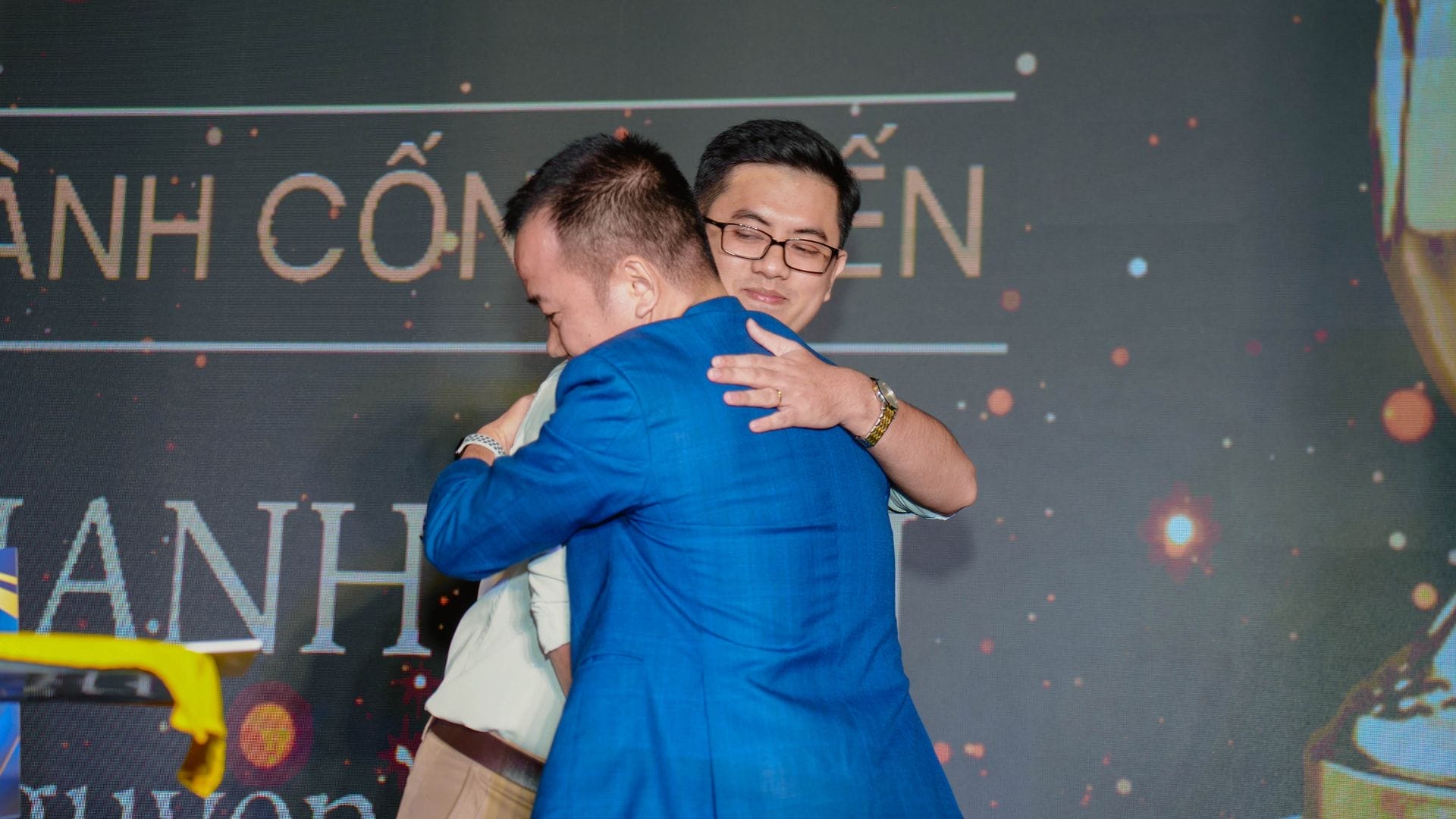 Two men are embracing on a stage, embodying the spirit of innovation and team building. One wears a blue suit and glasses, the other in a light-colored outfit. Behind them, a large screen bears partially readable text and vibrant lights, capturing the essence of an AXA Engineers event.
