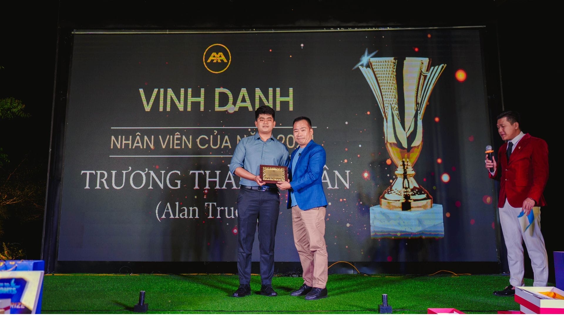 A man in a blue shirt receives a plaque from another man in a blue blazer at an AXA Engineers event. They stand on stage with a large screen displaying a trophy graphic, celebrating innovation. Nearby, another man in a red jacket looks on proudly.