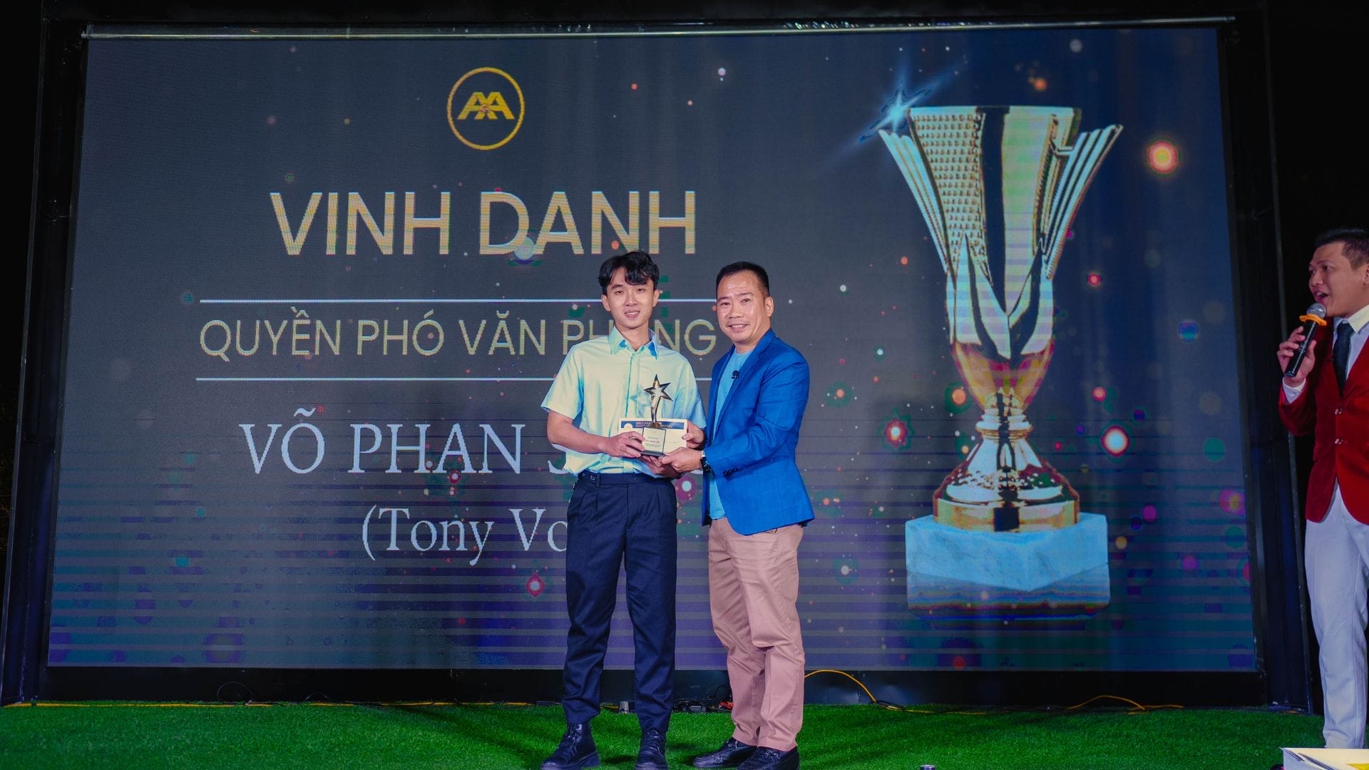 Two men stand on a stage during an AXA Engineers award ceremony. One holds a trophy while shaking hands with the other, celebrating their innovation. A large trophy image and text are displayed on the screen behind them.