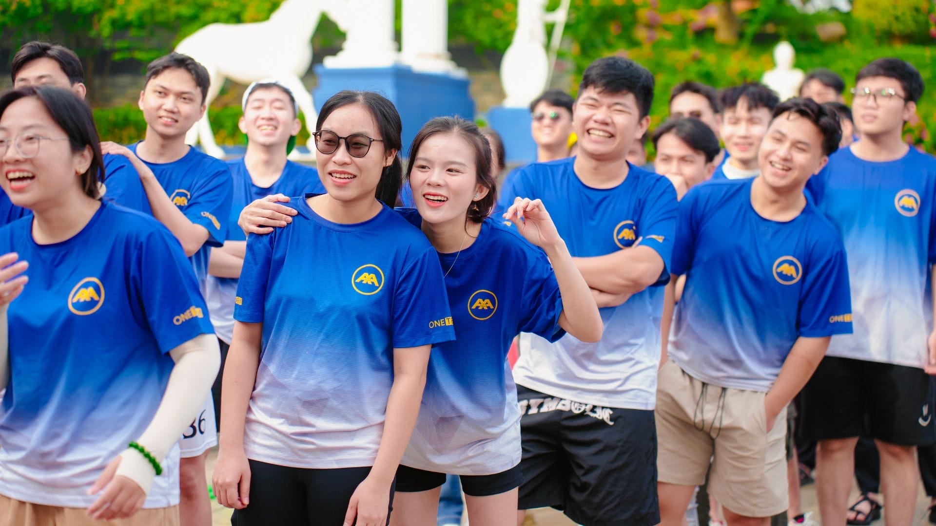 A group of smiling young adults stands in a sunny outdoor setting, wearing matching blue and white gradient T-shirts with a yellow logo. They seem to be participating in an AXA Engineers team-building activity, with trees visible in the background, fostering innovation among the team members.