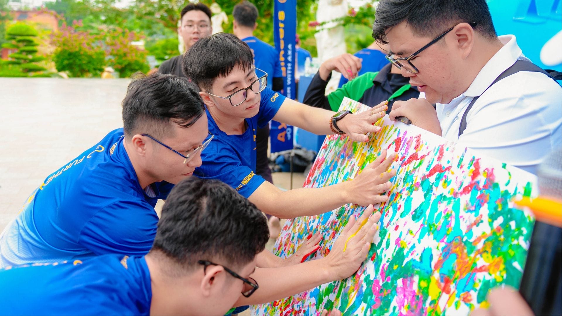 A group of people wearing blue shirts collaboratively works on a colorful abstract painting outdoors, embracing innovation. Engaged in this vibrant team-building activity, they focus on the artwork, using their hands to add touches of paint. The setting is bright and lively.