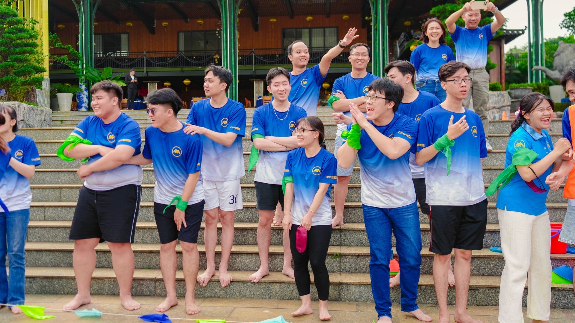 A group of people wearing matching blue shirts with white gradients stands and sits on outdoor steps. Embracing the spirit of innovation, some are smiling and waving, while others hold colorful objects. Its a lively and cheerful AXA Engineers 2024 atmosphere perfect for team building.
