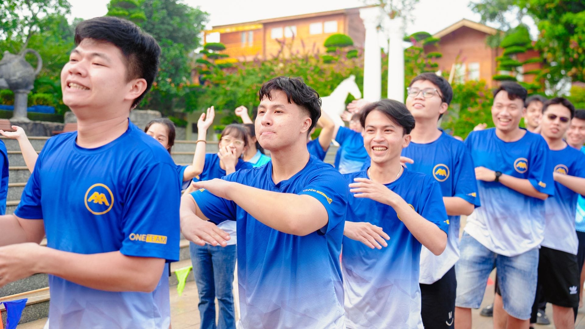 A group of people in matching blue and white shirts from the AXA Engineers 2024 team are standing outdoors in a line, smiling and enjoying an innovative team-building activity. They are in a garden setting with trees and a building in the background.