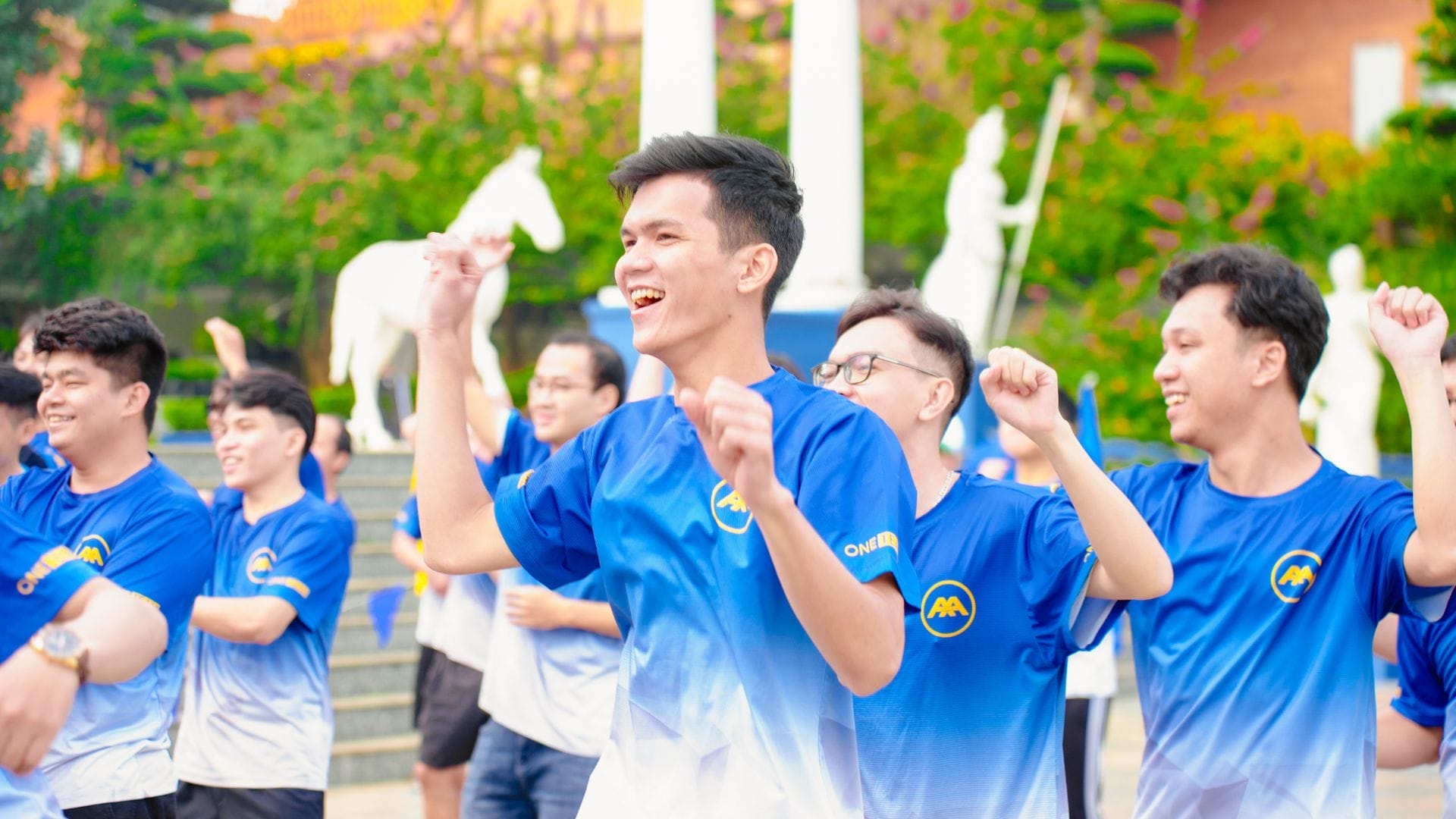 A group of young people in blue and white jerseys joyfully engage in team building, dancing outdoors amid statues and greenery. Raising their arms with smiles, they embody the spirit of innovation as they look forward to AXA Engineers 2024.