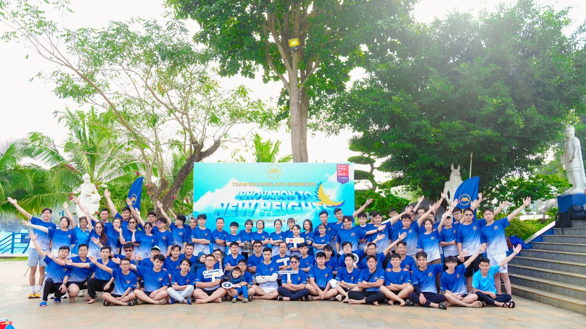 A large group of people wearing matching blue shirts pose cheerfully outdoors, radiating the spirit of team building. Gathered in front of a banner that reads Innovation to New Heights with vibrant trees in the background, they embody the excitement for AXA Engineers 2024.