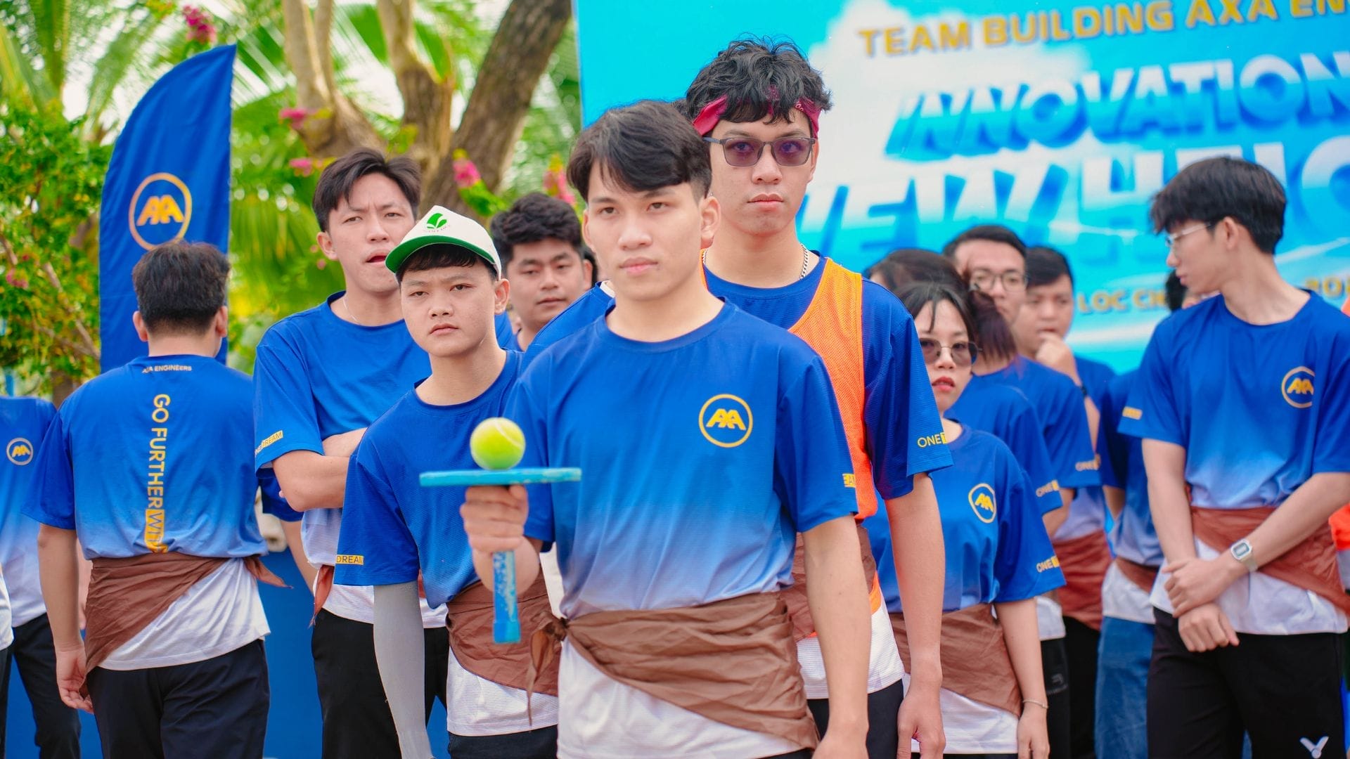A group of people in blue and white shirts engage in a lively team-building event. At the forefront, one person balances a ball on a spoon, showcasing their skill. A banner proudly displays Innovation at the AXA Engineers Team Building event.