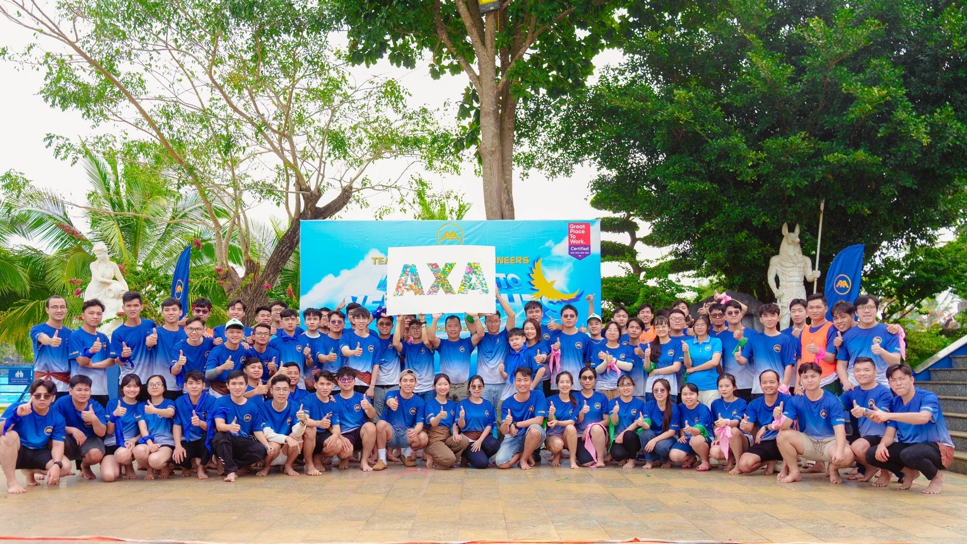 A large group of people wearing blue shirts, some barefoot, pose outdoors in front of a colorful backdrop with the AXA Engineers logo. Smiling and giving thumbs up, they celebrate innovation in team building. Trees and statues provide a charming background.