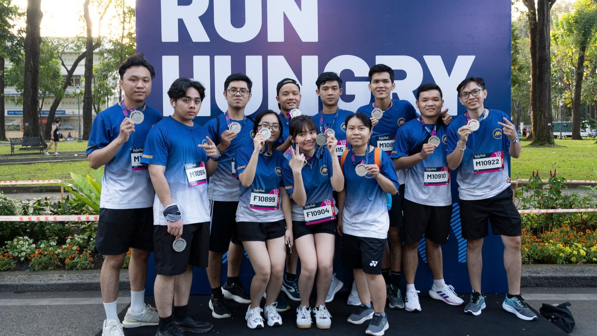 A group of people wearing blue and white athletic wear, representing AXA Engineers, pose proudly with medals in front of a Run Hungry sign. They are celebrating their achievement at the HCMC Marathon 2025, standing outdoors amid lush trees and vibrant flowers.