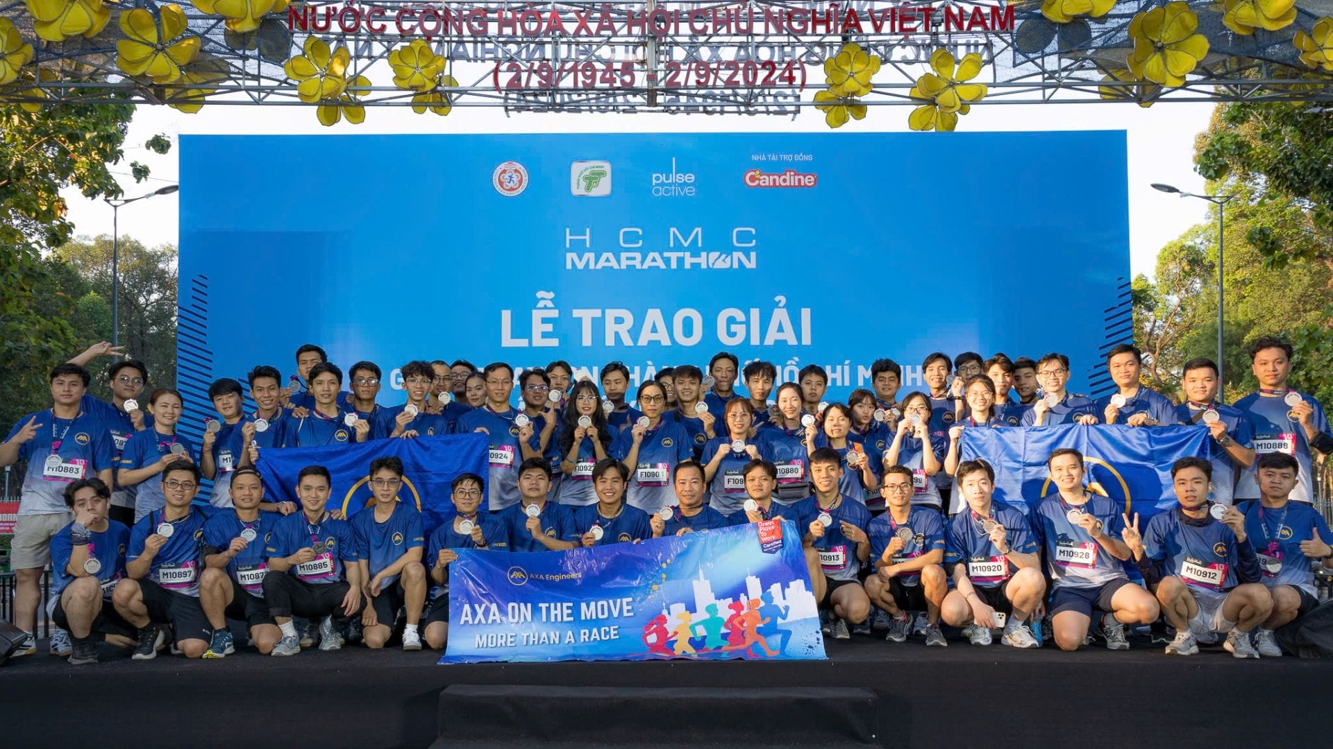 A large group in matching blue attire proudly pose with banners at the outdoor HCMC Marathon 2025 event, conquering the challenge before a blue stage backdrop displaying various logos. Trees and decorations add charm to this vibrant scene.