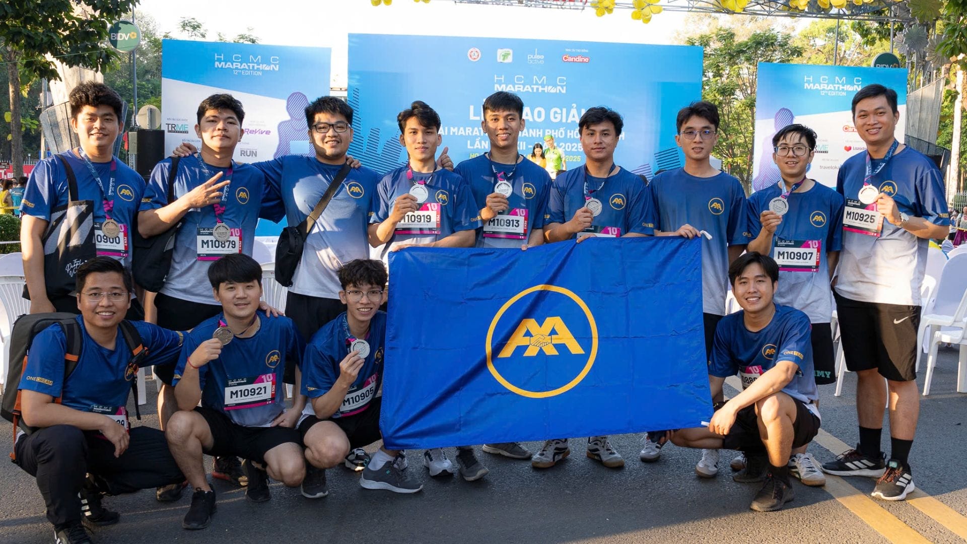 A group of smiling individuals wearing matching blue shirts poses together outdoors, proudly holding a blue flag with a yellow emblem. Bearing marathon bibs, they stand under a banner that reads H.C.M.C Marathon 2025. Among them are members of the dedicated AXA Engineers team.