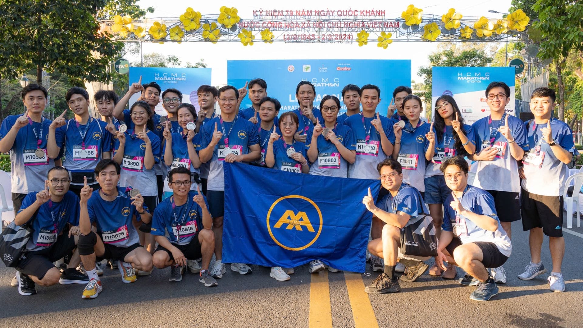 A group of people in matching blue shirts pose together, proudly holding a blue flag with a yellow logo. They are outdoors under a decorative arch with flowers, embodying the spirit of the AXA Engineers at the HCMC Marathon 2025, their race numbers prominently displayed on their shirts.