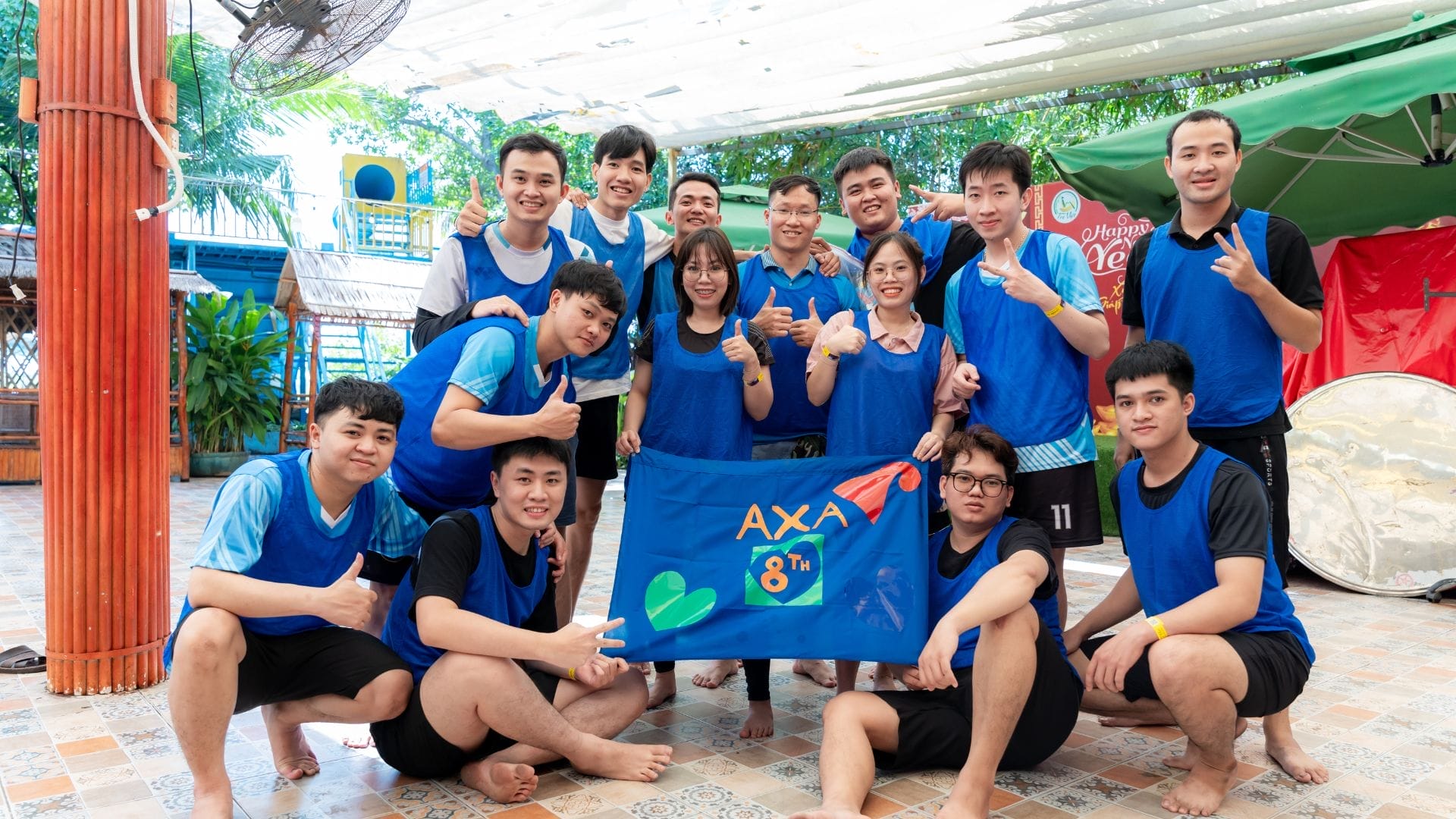A group of 15 people wearing matching blue sports jerseys smile and pose together, embodying a shining success. Theyre holding a vibrant blue banner adorned with colorful decorations. The scene unfolds in a charming outdoor area with trees and a covered patio, celebrating the AXA Engineers Buddy Game 2024.