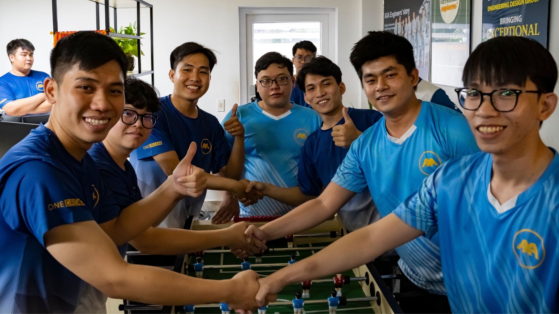 A group of eight young men stands around a foosball table, smiling and giving thumbs up, embodying the spirit of connection through the AXA Engineers Buddy Game. They wear matching blue shirts, some with glasses, against a backdrop of posters and a door.