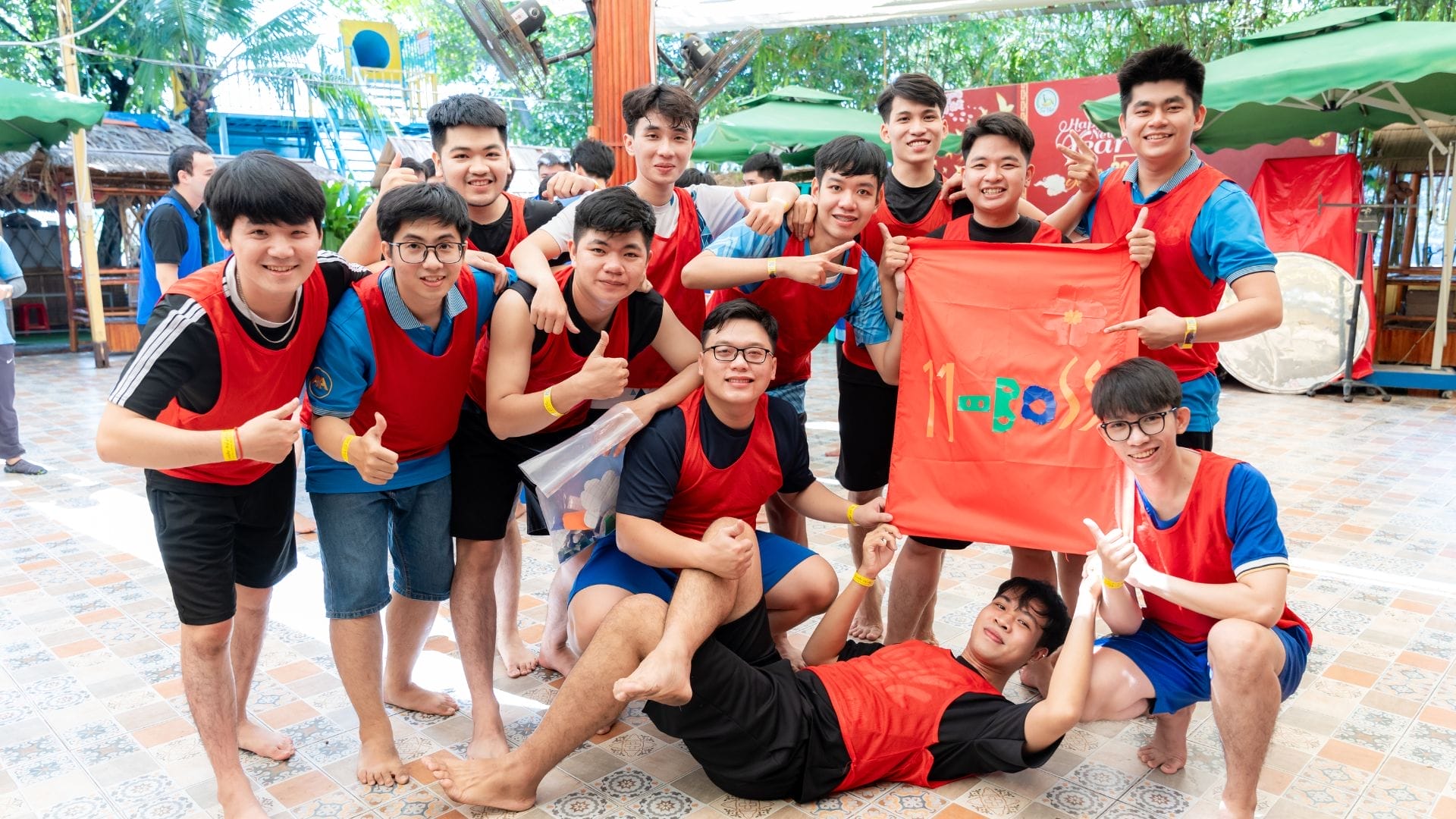 A group of young people, mostly men, in matching red and blue athletic vests smile for a photo during the AXA Engineers Buddy Game. Holding a vibrant red flag with colorful letters, they stand on patterned tiles with greenery and tents behind them, celebrating connection and success outdoors.