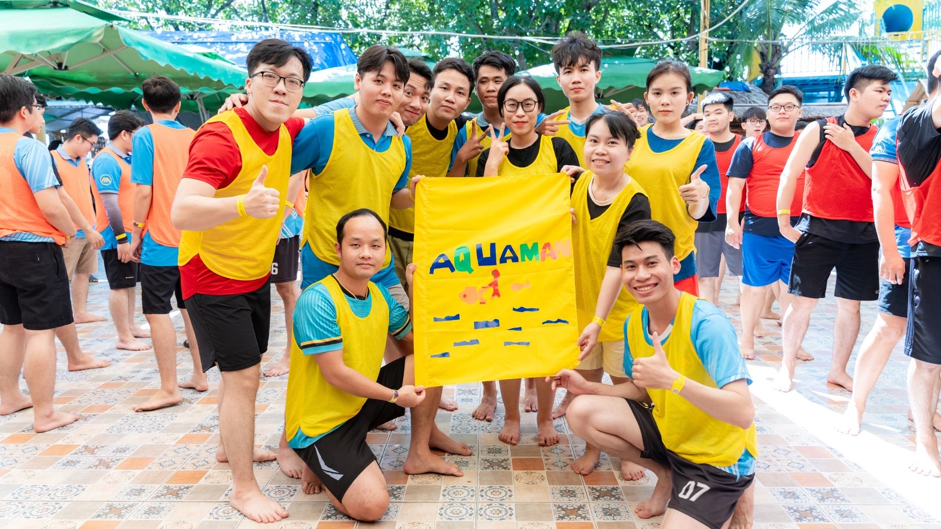A group of people wearing yellow jerseys stand outdoors, smiling and holding a yellow banner with the word AQUAMAN on it, embodying the spirit of connection. Others in blue and red jerseys are in the background, enjoying what seems to be an AXA Engineers Buddy Game or sports day celebration.