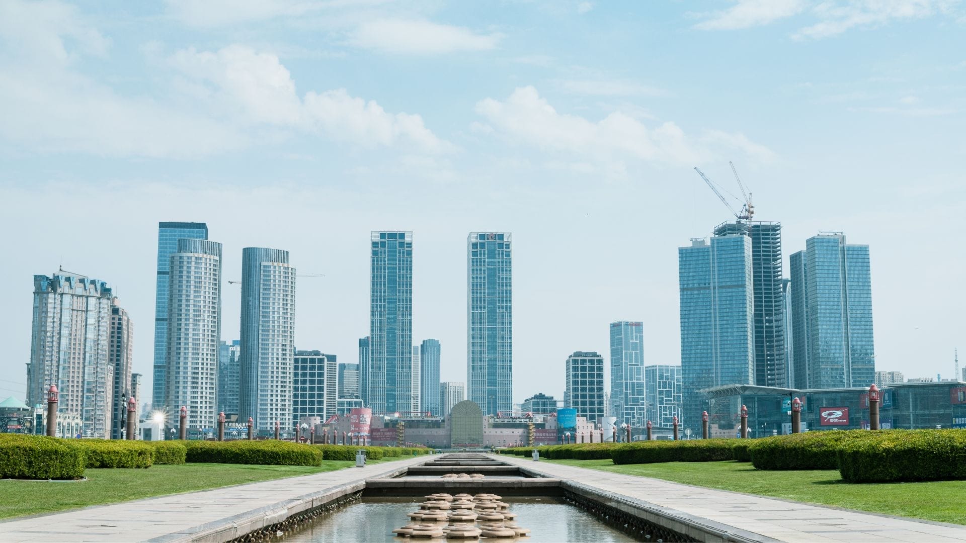 A modern cityscape highlighting tall skyscrapers under a clear blue sky. In the foreground, a landscaped park features a long, narrow reflecting pool with circular stepping stones, flanked by manicured hedges—an embodiment of future trends in civil engineering.