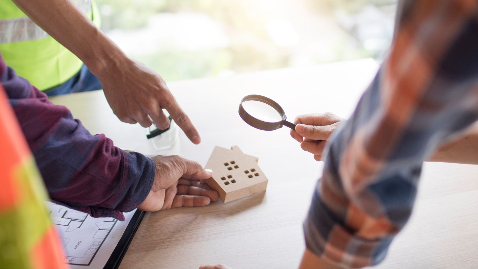 Three people examine a small wooden house model on a table. One person holds a magnifying glass while another points at the house. A blueprint is partially visible as they discuss scaling up civil engineering projects, focusing on construction and architecture details.