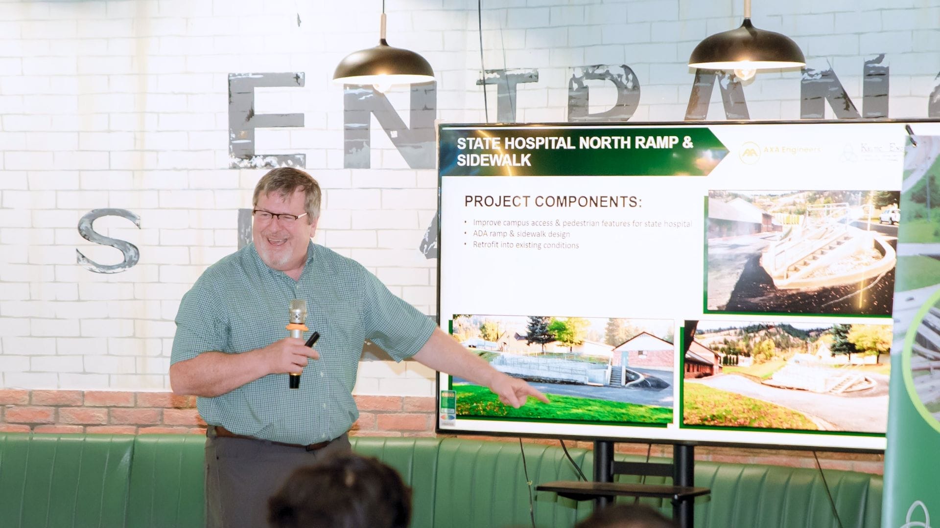 A man in a green shirt from AXA Engineers is speaking into a microphone and pointing at an opportunity on a presentation slide titled State Hospital North Ramp & Sidewalk, which displays images and text on project components.