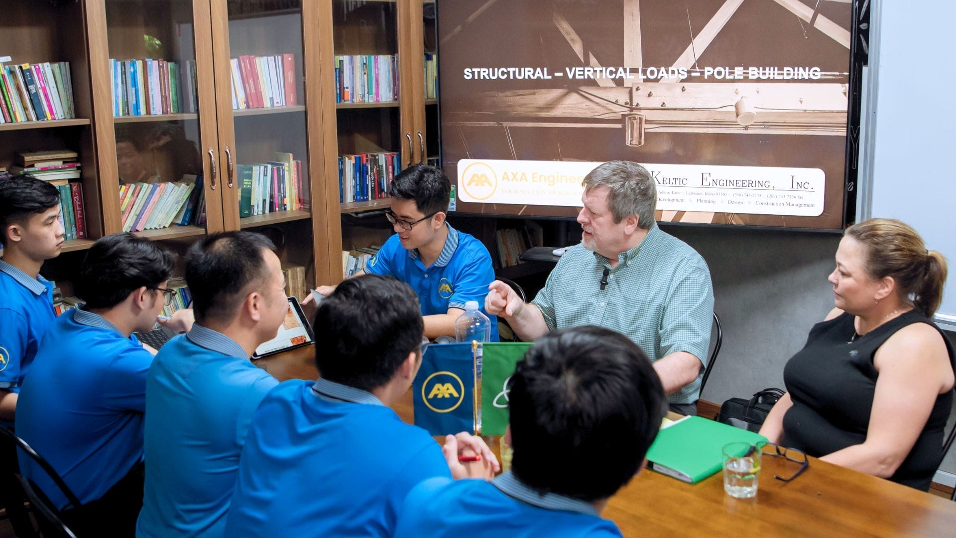 A group of people in blue shirts sit around a conference table, attentively listening to a man and woman from AXA Engineers at the head. A presentation on structural engineering and pole building offers an opportunity for PE certification, while bookshelves line the room.