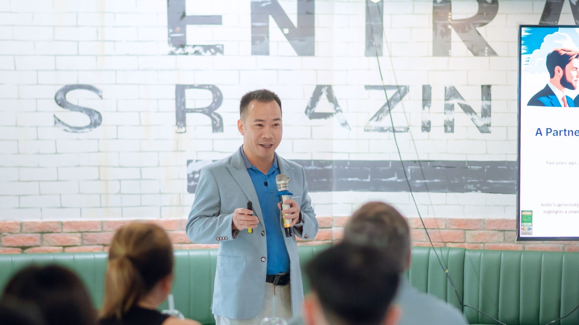 A person in a blue blazer is speaking into a microphone at the Welcome Meeting 2025, engaging a small audience. They stand near a screen displaying Partnership as they discuss opportunities. The backdrop of the white brick wall features partial lettering, setting an inspiring scene.