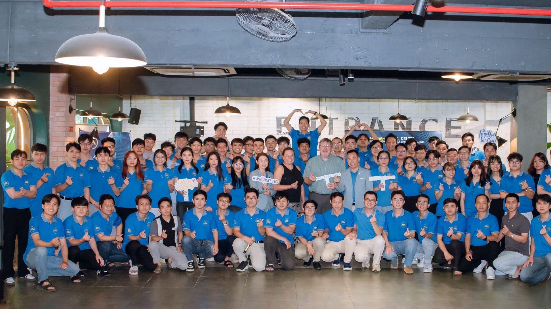 A large group of about ninety people wearing blue shirts pose together under industrial-style lighting. Signs highlight Welcome Meeting 2025, while some give thumbs-up gestures, capturing the excitement for new learning and development opportunities. Smiles beam at the camera.