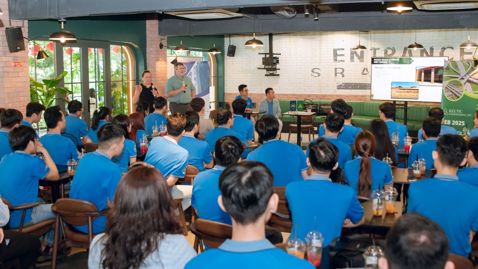 A group of people in blue shirts are attending a Welcome Meeting 2025 presentation in a modern, casual indoor setting. Two presenters stand at the front near a screen displaying slides. Attendees are seated, listening attentively to potential opportunities being discussed.