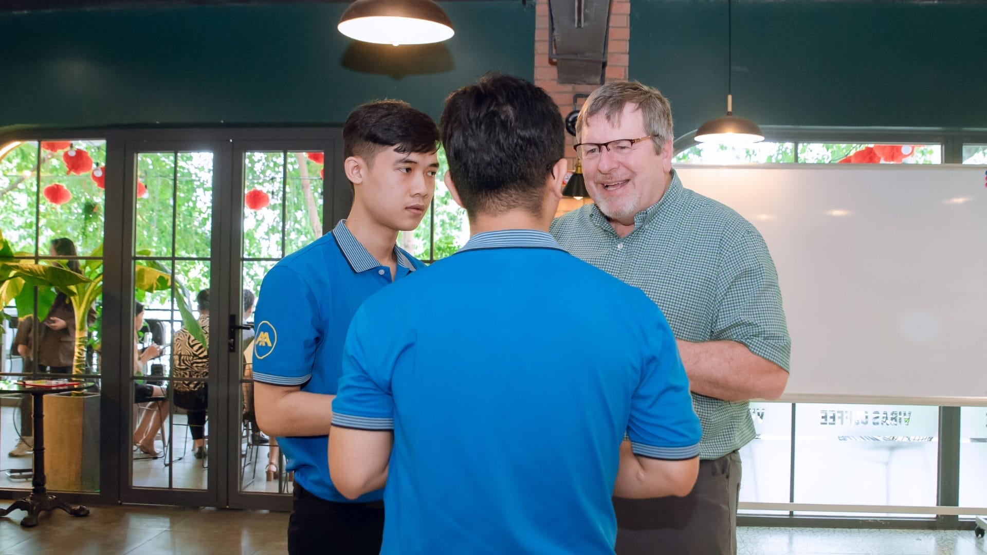 A man in a green shirt is engaging in conversation with two younger men wearing blue uniforms in a brightly lit room with glass windows. They stand near a glass door, discussing opportunities for learning and development, with plants visible outside, setting the tone for the Welcome Meeting 2025.