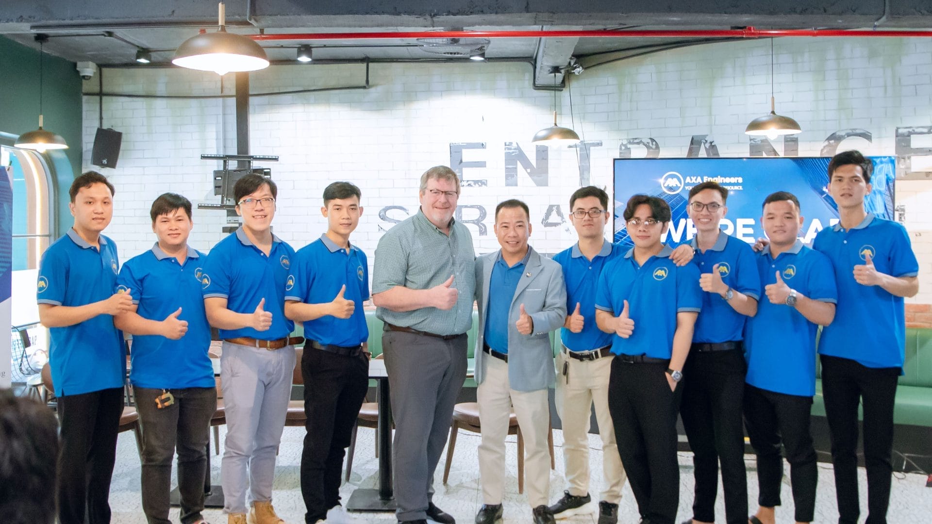 A group of people, mostly in blue shirts, smiling and giving thumbs up at the 2025 Welcome Meeting. Theyre posing for a photo in front of a backdrop with text and logos, set against a room with industrial-style decor.