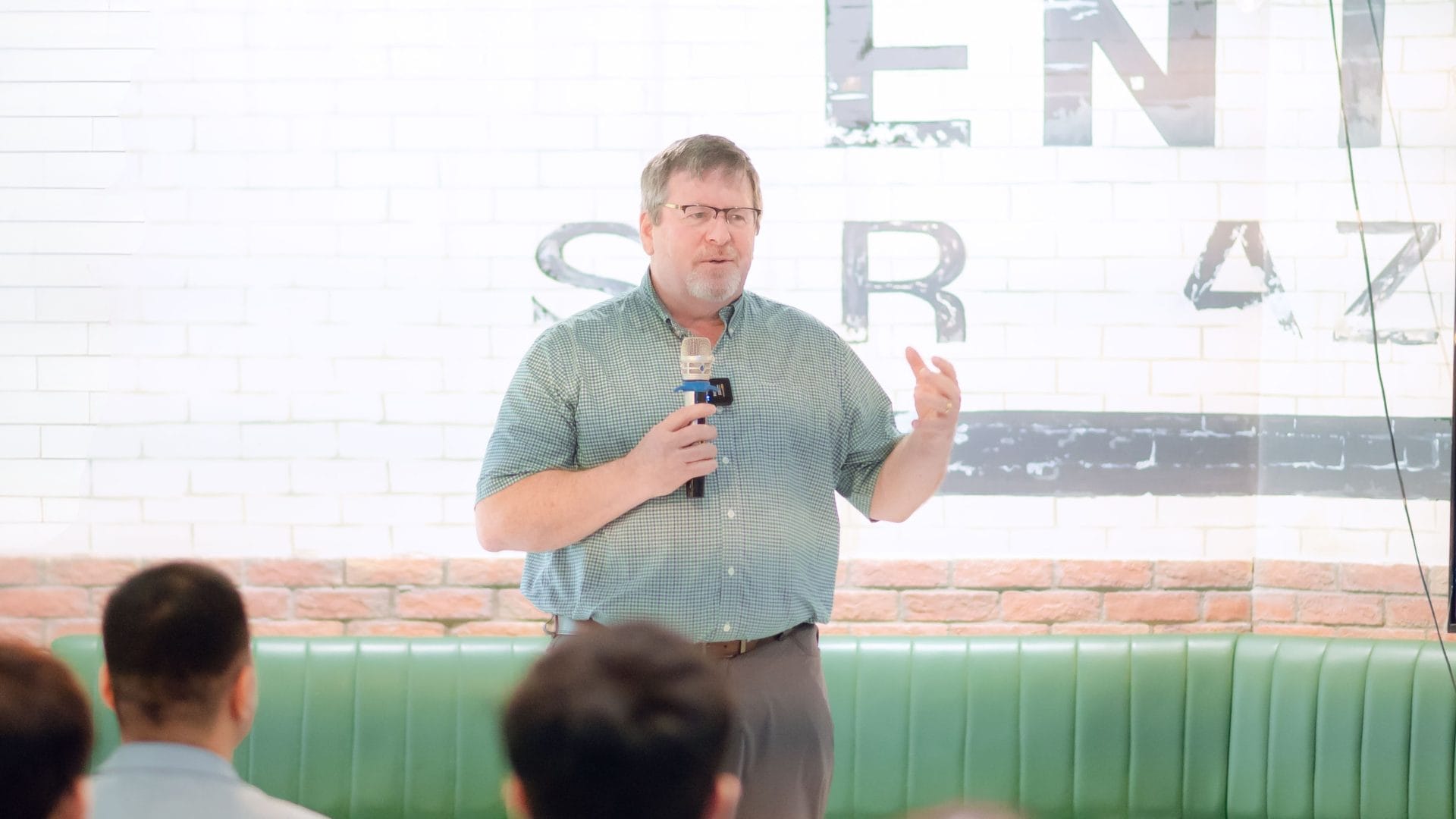A man in a green shirt is speaking into a microphone in front of an audience, highlighting opportunities for growth. He stands against a white brick wall with partial text visible, suggesting the backdrop for the Welcome Meeting 2025 in this casual indoor venue.