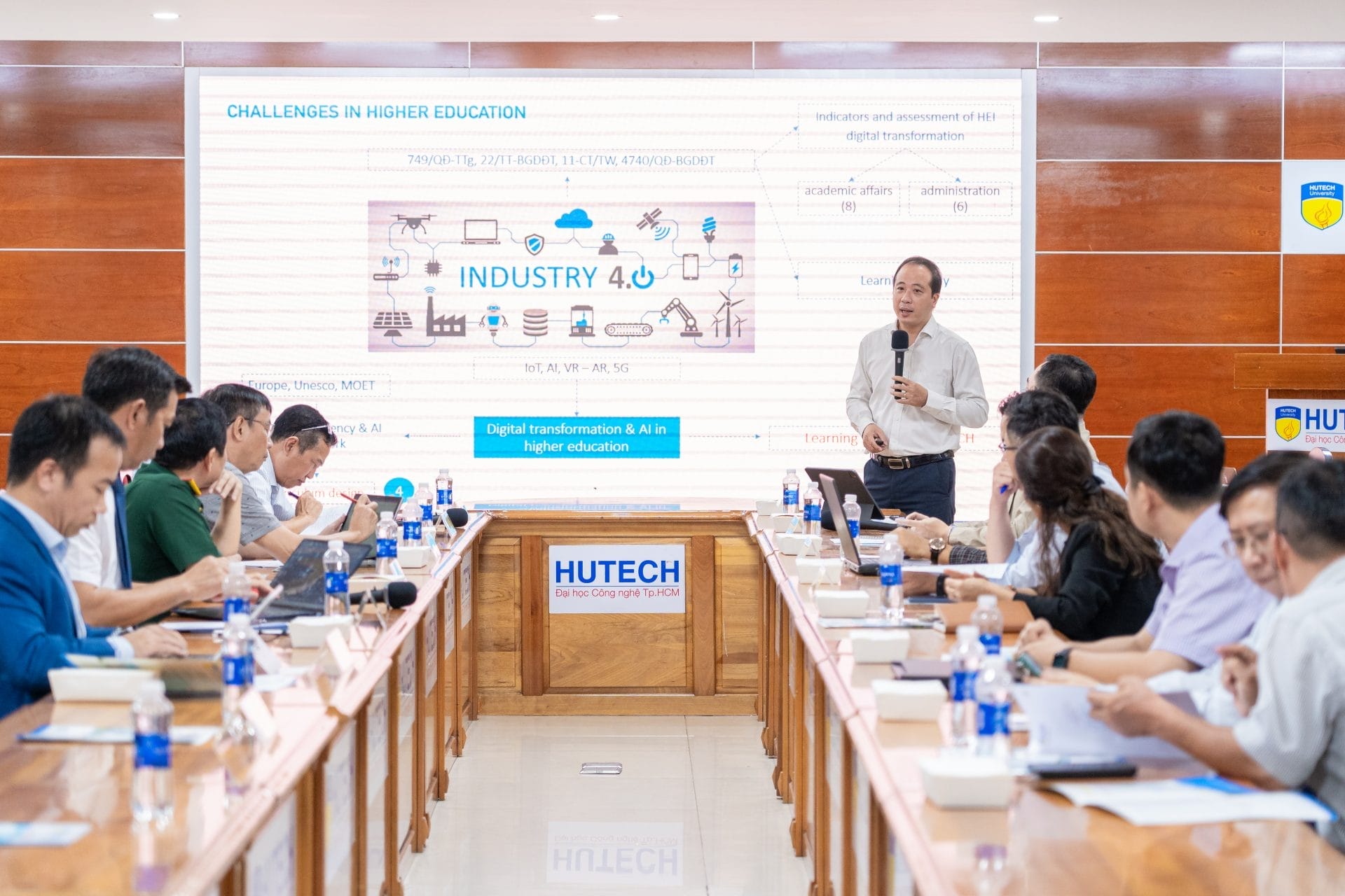 A man stands at the front of a conference room presenting a slide titled Challenges in Higher Education with an emphasis on finding motivation amidst Industry 4.0. Attendees, including students, seated at a long table, listen and take notes in the wooden-walled room.