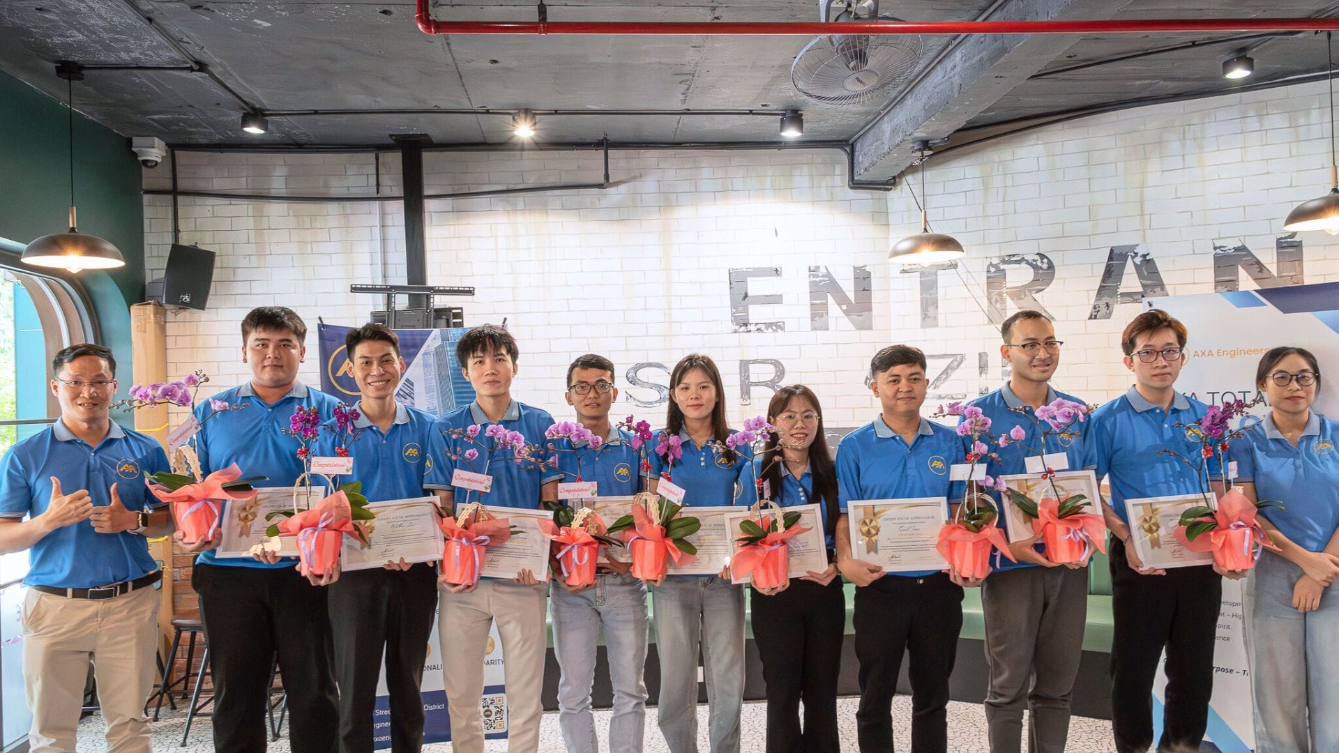 Civil Engineering Outsourcing A group of eleven individuals, all dressed in blue shirts, stands in a modern, industrial-style room brightly lit indoors, holding certificates and potted purple orchids. They smile at the camera following an ATED seminar that emphasized holistic development and the latest trends in civil engineering outsourcing.
